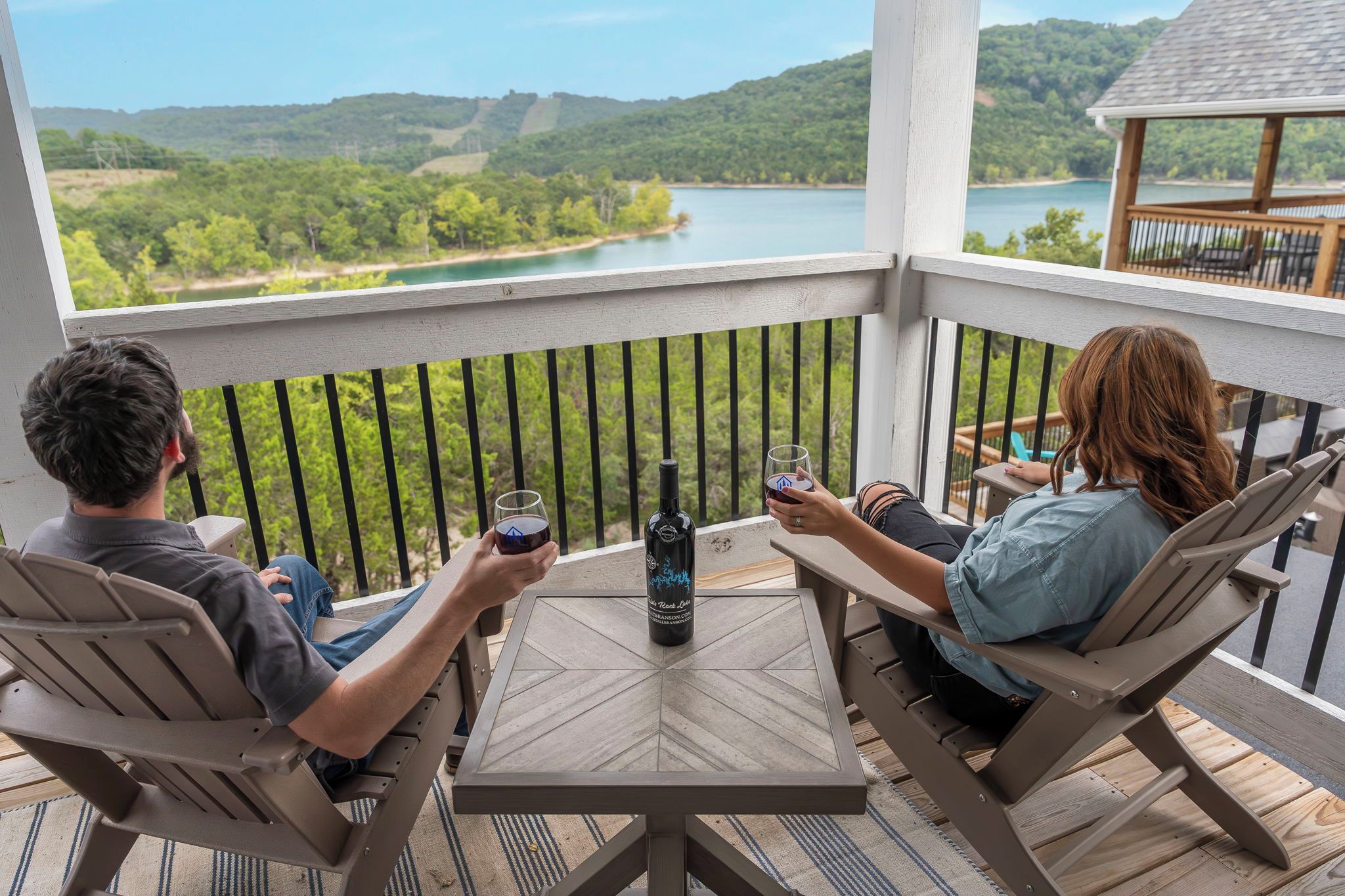 A couple seated at a table on a balcony overlooking a lake, with a bottle of wine on the table.