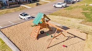 Ein Spielplatz mit einer hölzernen Spielkonstruktion, darunter Schaukeln und Kletterelemente, umgeben von Sand.
