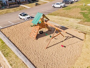 Ein Spielplatz mit einer hölzernen Spielkonstruktion, darunter Schaukeln und Kletterelemente, umgeben von Sand.