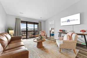 A modern living room with a leather sofa, a striped armchair, and a coffee table. A large window offers a view of a balcony. - Condo in Kimberling City w Lake View_Cmty Pools_Free Attaction Tickets (Kimberling City)