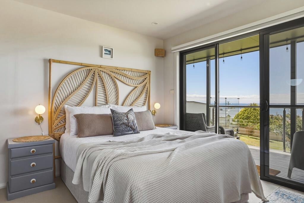 A bedroom with a large bed, a nightstand, a wall-mounted lamp, and a view through a sliding glass door.
