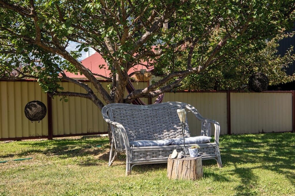 A wicker outdoor seating area with a bench and a small table, surrounded by a fence and a tree.