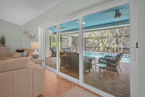 A living room with a beige sofa, a coffee table, and a lamp. There is a sliding glass door leading to an outdoor patio with a pool and lounge chairs. - Bonita 3BR | Pool | Coconut Jack’s 3mi (Bonita Springs)