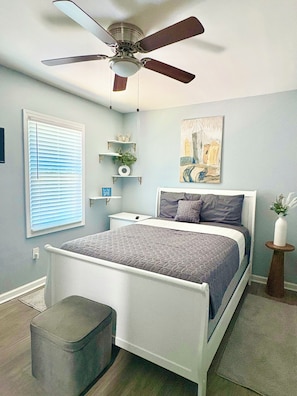A bedroom with a bed, a ceiling fan, a window with blinds, a bedside table, a nightstand, a wall-mounted shelf with decorative items, and a painting. - Gorgeous 3-bedroom bungalow with AC in vibrant Atlanta (Atlanta)
