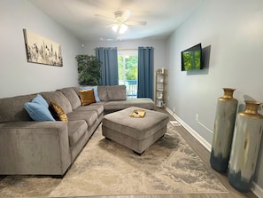 A living room with a sectional sofa, an ottoman, a ceiling fan, a television, and a large window with curtains. - Gorgeous 3-bedroom bungalow with AC in vibrant Atlanta (Atlanta)
