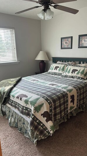 A bedroom with a bed, a ceiling fan, a window with blinds, a lamp, and two framed pictures on the wall.