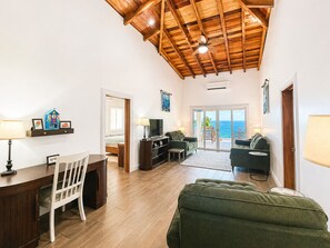 A spacious living area with a wooden ceiling, a television, a sofa, a desk with a chair, and a view of the ocean through a sliding glass door. - Family friendly beach front two-bedroom home with AC (Hopkins)