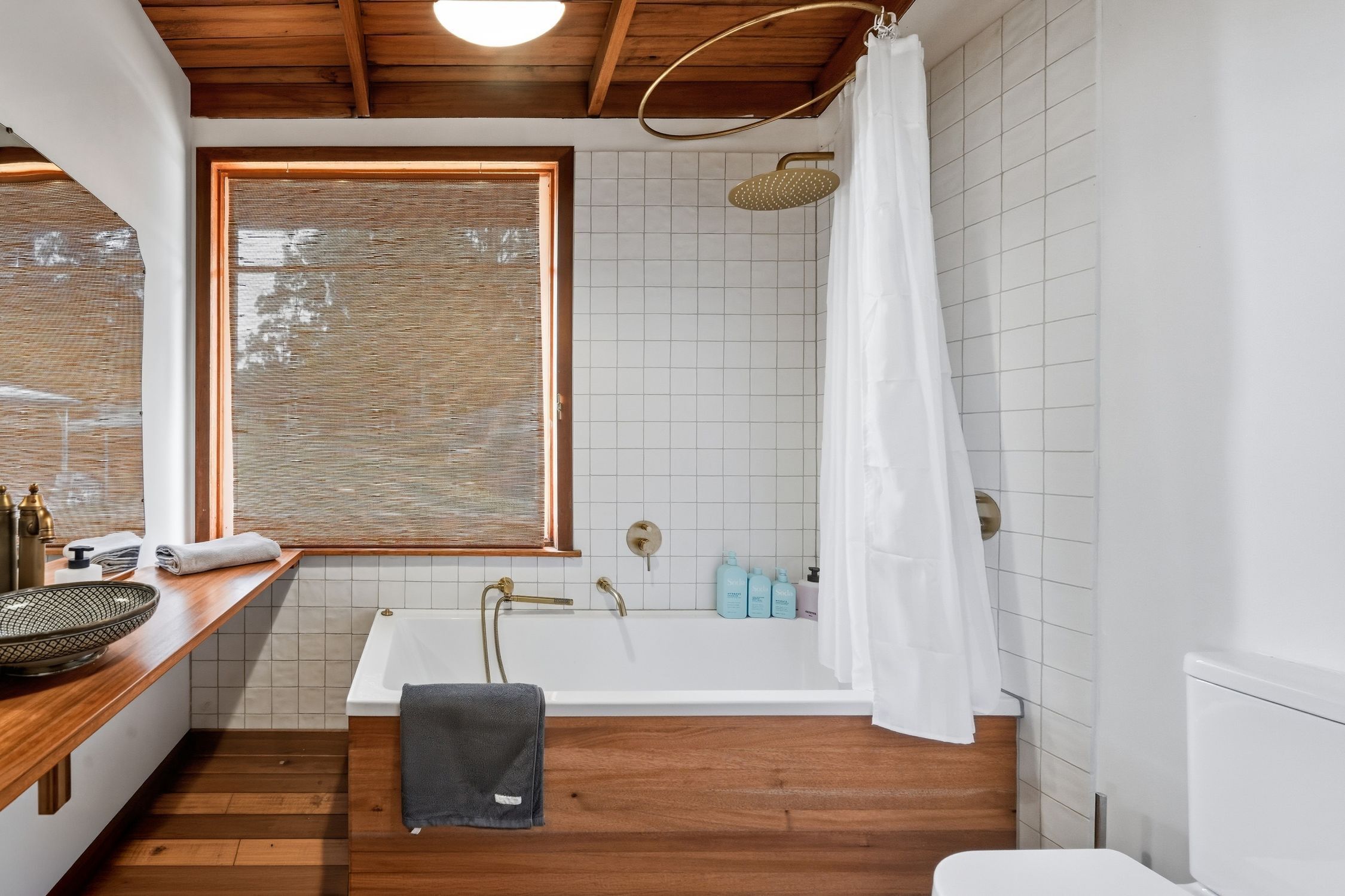 Une salle de bain avec une baignoire, une douche, une fenêtre avec des stores et un plancher en bois.