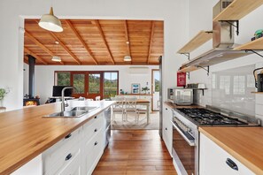A modern kitchen with wooden beams, a central island, and a view of a forested landscape. - Crofters Rest: Light Filled Hideaway on Tasman Peninsula (152 Robinsons Rd)