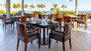 A restaurant with a view of the ocean, featuring round tables and chairs, and a tropical outdoor setting. - Luxurious oceanfront 2-bedroom suite located on pristine beach in Cabo San Lucas (San José del Cabo)