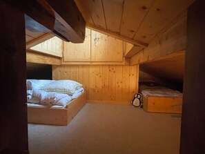 Une petite pièce en bois, avec un plafond en pente, un lit simple recouvert d’une courtepointe à motifs blancs et verts, une petite table de chevet et une horloge fixée au mur.