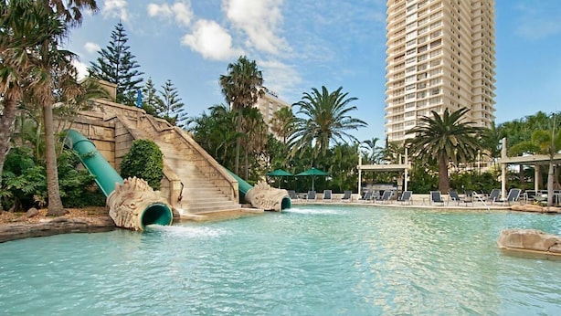 Una piscina con cascada, sillas de descanso, palmeras y un edificio de hotel alto al fondo.
