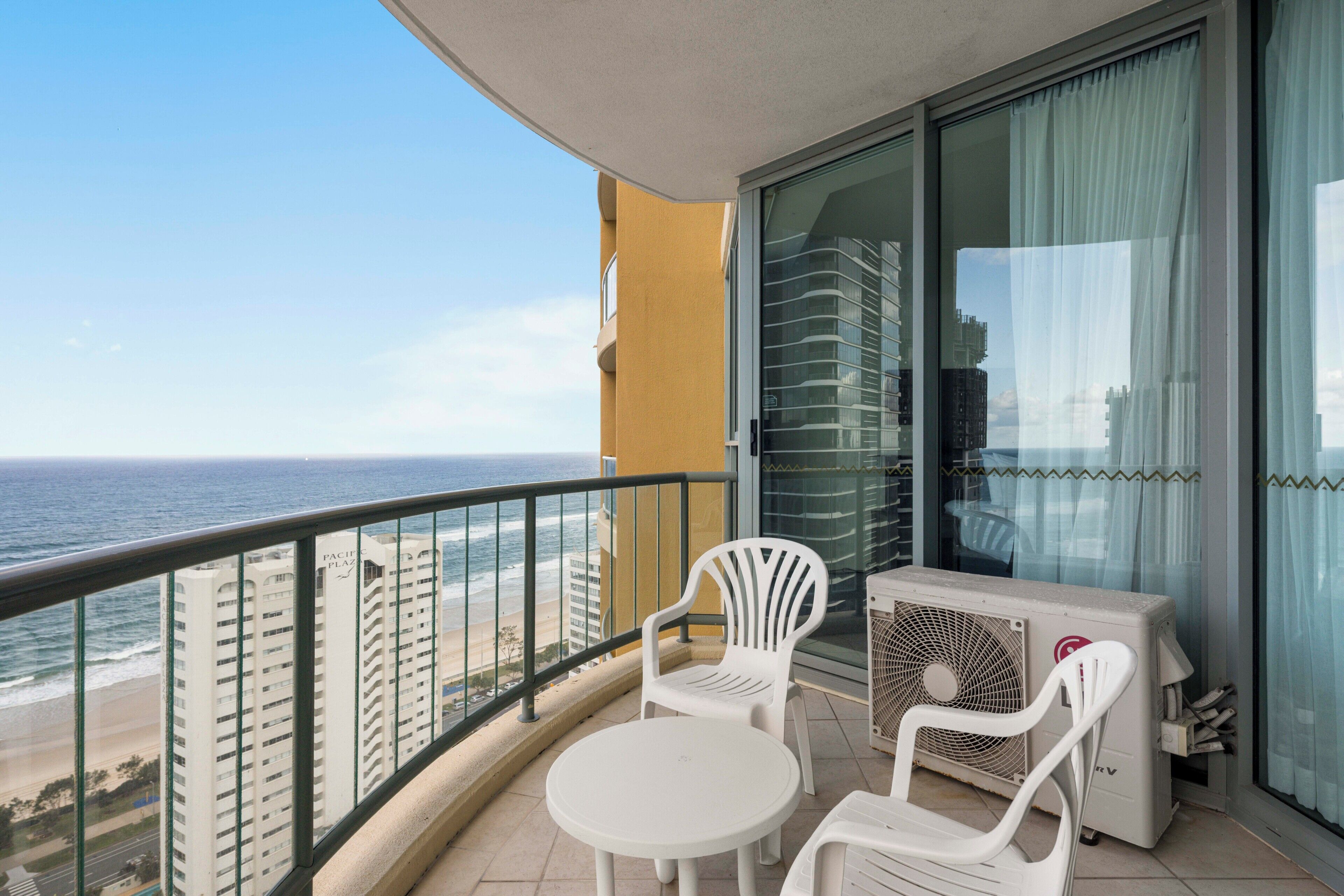 A balcony with a view of the ocean, a white table, two white chairs, and an air conditioning unit.