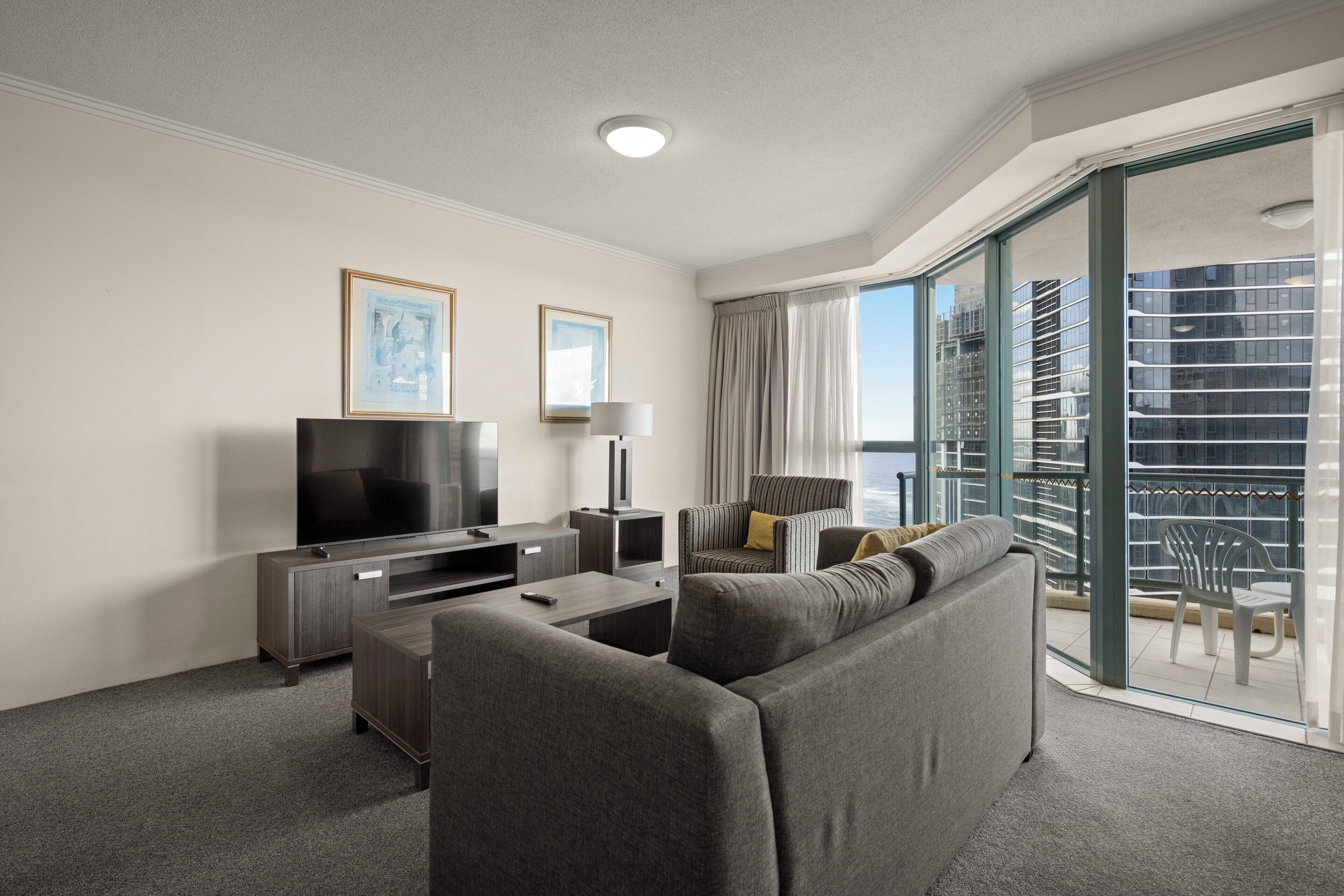 Habitación de hotel moderna con sofá gris, televisor de pantalla plana, una mesita y balcón con vistas a la ciudad.