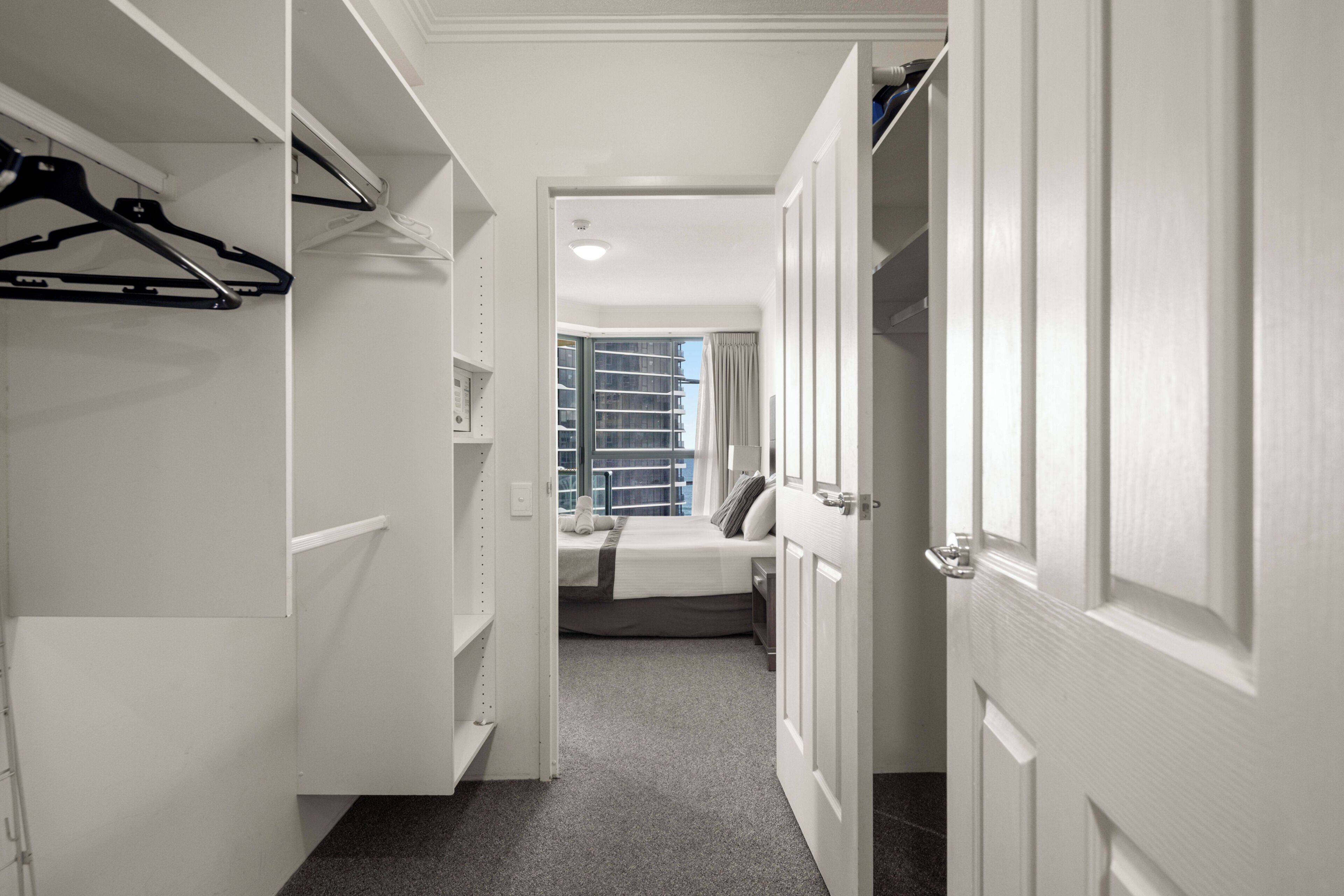 A hallway leading to a bedroom with a bed, a window, and a closet with hanging clothes.