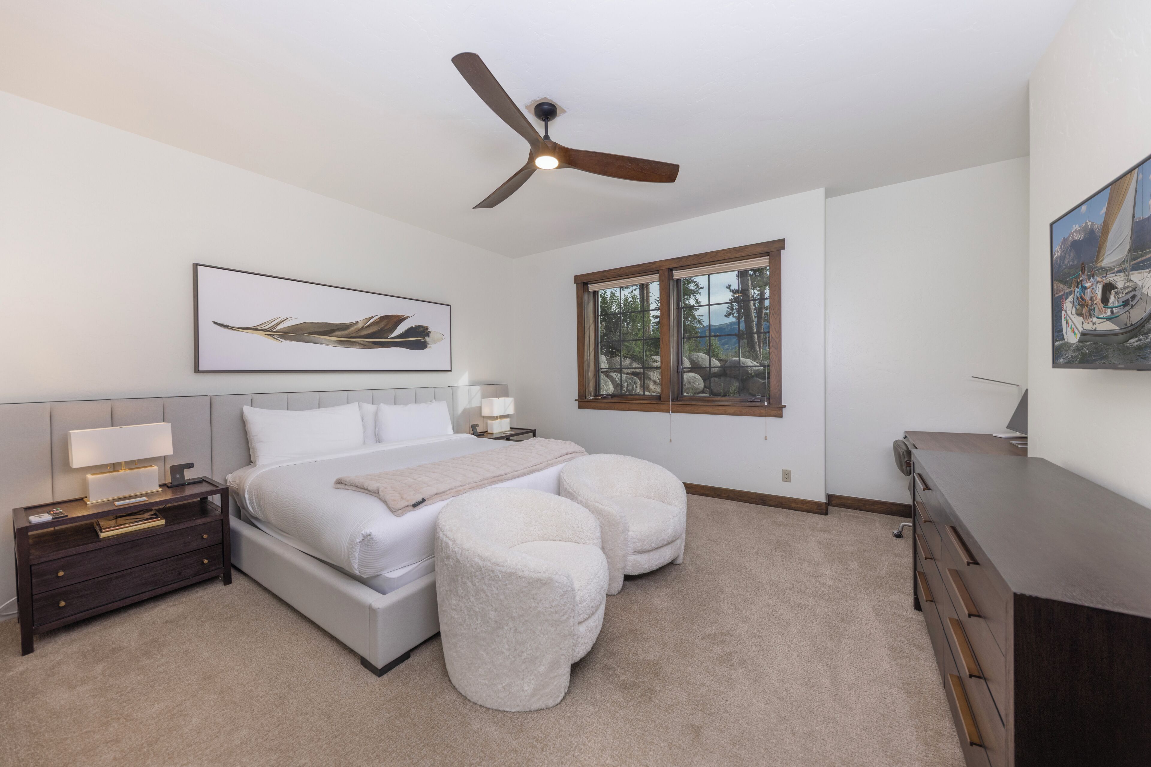 A bedroom with a bed, nightstand, two ottomans, a ceiling fan, and a window with a view.