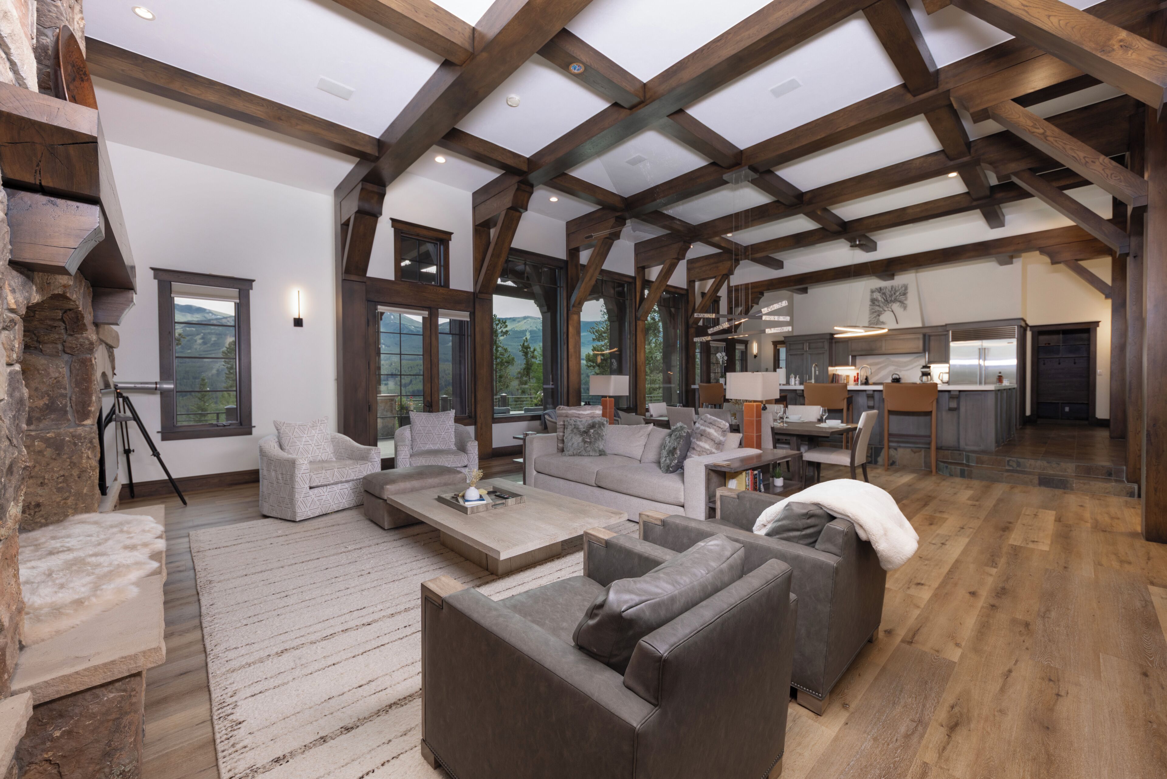 A spacious living area with a high ceiling and exposed wooden beams, a stone fireplace, and large windows offering a view of the outdoors.