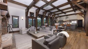 A spacious living area with a high ceiling and exposed wooden beams, a stone fireplace, and large windows offering a view of the outdoors.