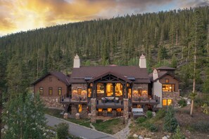 A large, two-story lodge with a stone facade, multiple chimneys, and a spacious porch with outdoor seating. - Unmatched Elegance, Pool Table, Hot Tub, Ski Shuttle, Fire PIt with Views!! (Breckenridge)