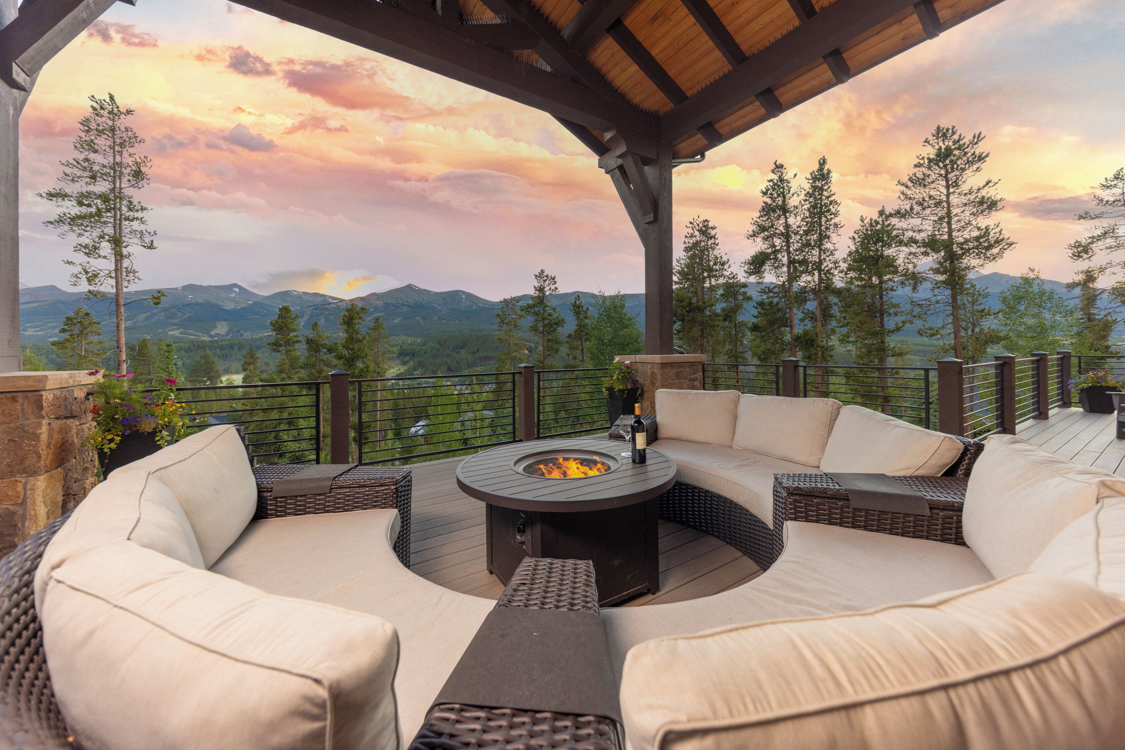 An outdoor patio with a round fire pit table, cushioned seating, and a view of mountains.