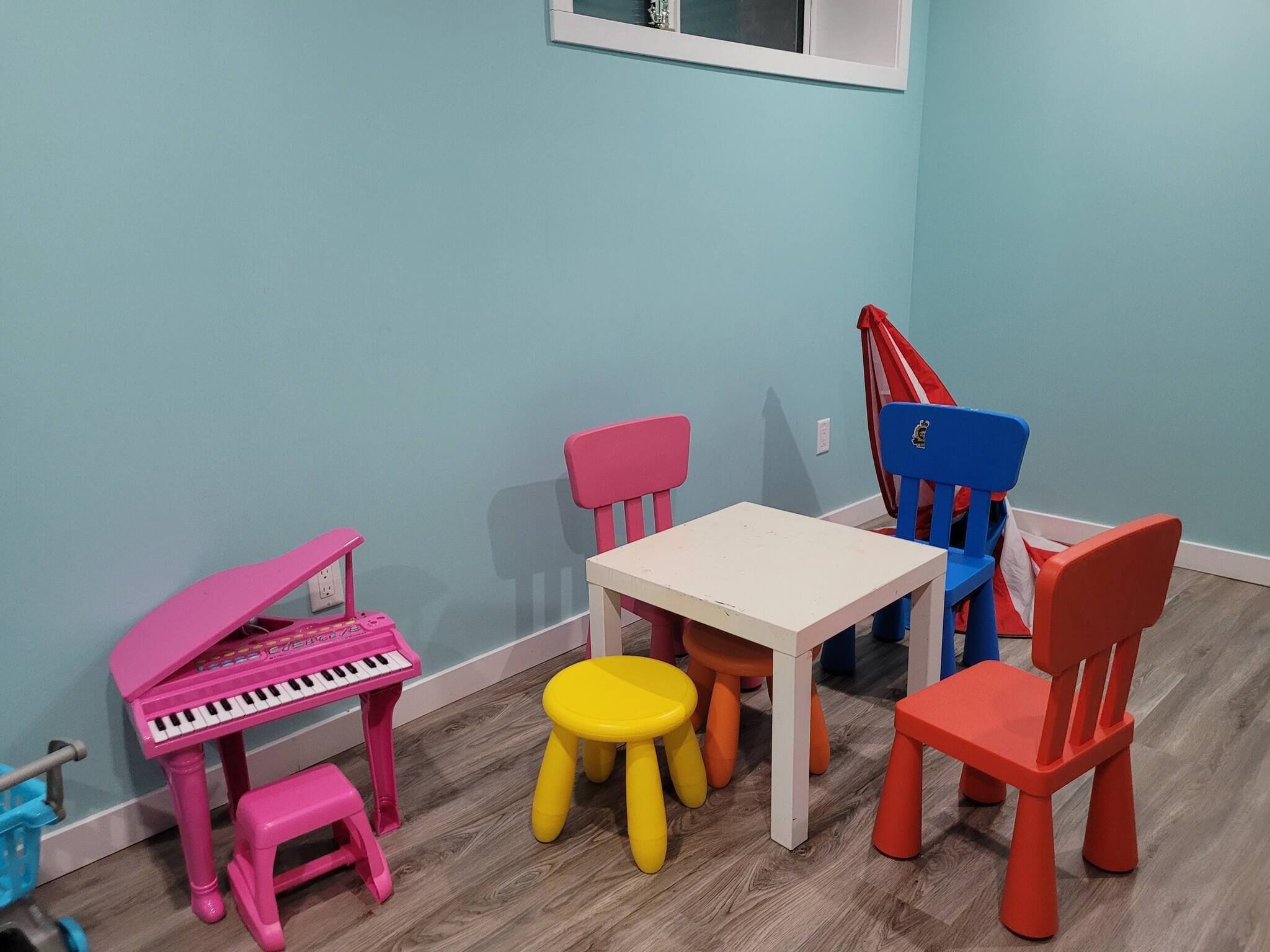 A children's play area with a pink piano, a white table, and colorful chairs.