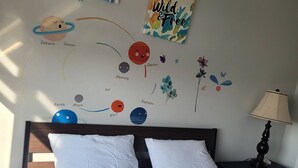 A bedroom with a bed, pillows, a nightstand, a lamp, and wall art depicting planets and flowers.