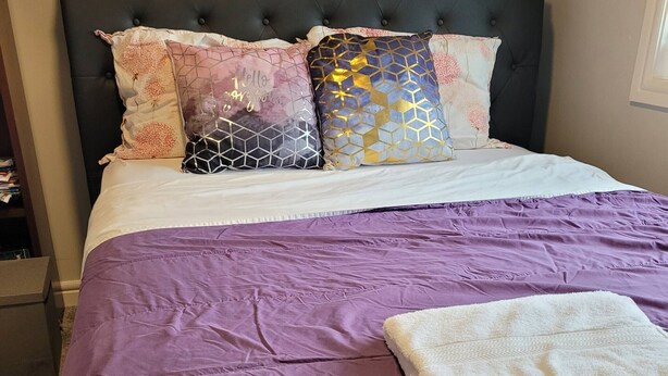 A bed with a purple bedspread, a white blanket, and multiple decorative pillows.