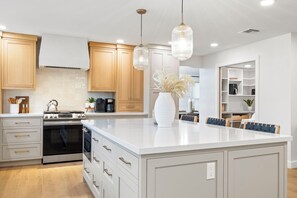 Uma cozinha moderna com armários brancos, uma ilha grande, eletrodomésticos de aço inoxidável e iluminação pendente.