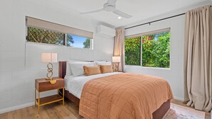 Un dormitorio con cama, mesitas de noche, ventilador de techo y ventana con vistas a follaje.