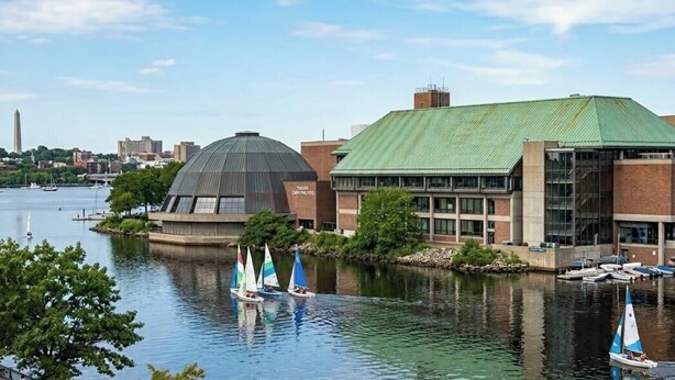 A waterfront building with a green roof, sailboats on the water, and a city skyline in the background. - Brand New Spacious Family Friendly Home near Boston (Somerville)