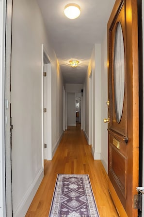 A hallway with wooden flooring, a patterned rug, a wooden door with a glass panel, and a ceiling light fixture. - Brand New Spacious Family Friendly Home near Boston (Somerville)