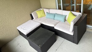Ein Ecksofa mit Kissen, ein kleiner Hocker und ein Korbstuhl auf einem Balkon mit strukturierter Wand und Glastür.