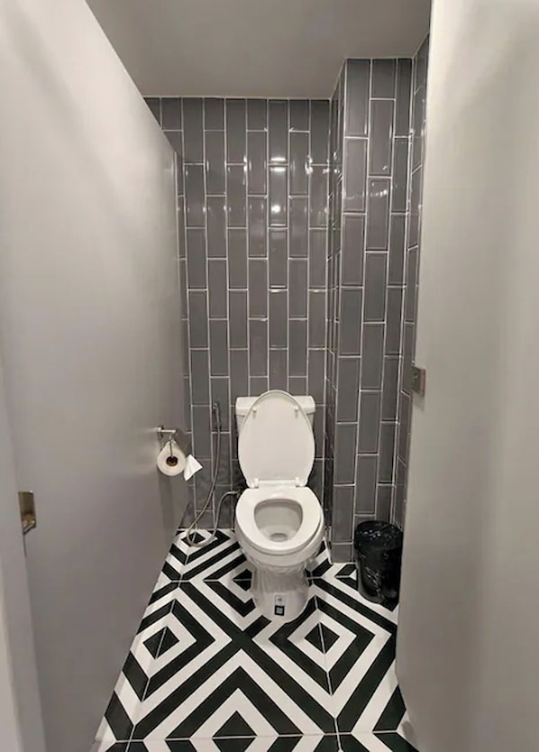 A bathroom with a white toilet, gray tiled walls, and a black and white patterned floor. - The Rich Cafe & Hostel (Pattaya)