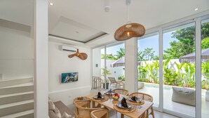 Un comedor con una mesa dispuesta para cuatro, rodeado de sillas de mimbre, un ventanal con vistas a zonas verdes y una escalera que sube a un nivel superior.