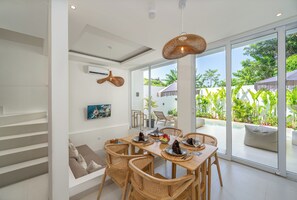 Un comedor con una mesa dispuesta para cuatro, rodeado de sillas de mimbre, un ventanal con vistas a zonas verdes y una escalera que sube a un nivel superior.