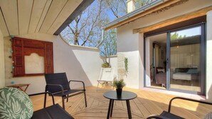 A covered outdoor area with a wooden ceiling, a seating arrangement with chairs and a table, a sliding glass door leading to a bedroom, and a potted plant.