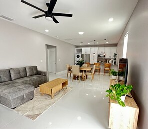 A modern living room with a sofa, coffee table, dining area, kitchen, ceiling fan, and potted plants.