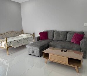 A modern living room with a grey sectional sofa, a matching ottoman, a small wooden coffee table, and a metal-framed bed with a white bedspread.