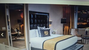 A hotel room with a large bed, a bench, a desk, and a view of the city at night.