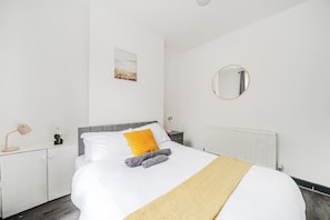 Un lit bien fait, recouvert d’une couette blanche, avec une seule serviette grise et un plaid de couleur moutarde. Un miroir rond est accroché au mur, au-dessus du lit.