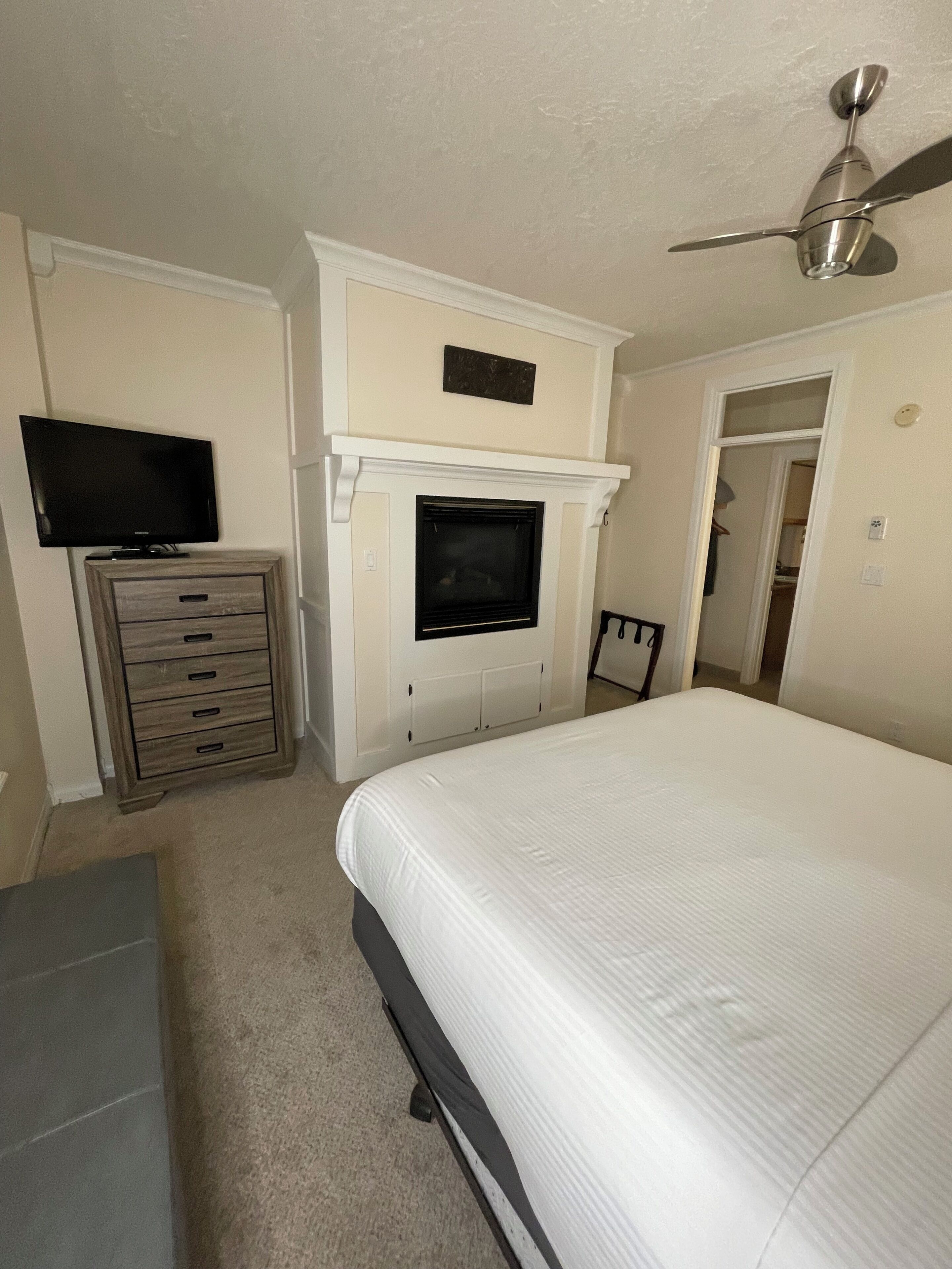 Ein Schlafzimmer mit einem Bett, einem Fernseher auf einer Kommode, einem Deckenventilator und einem Kamin.