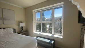 A bedroom with a large window, a bed, a bedside table with a lamp, and a television on a dresser. - Ski-Lift Next Door, 6 Guest, 2BR 2BA Pool & Spa, Sauna, Gym, Restaurant, Wi-Fi (Brian Head)