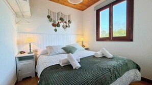 A neatly made bed with a green blanket, two bedside lamps, a wall hanging, and a window with a view of trees.