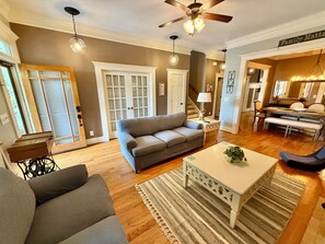 A living room with a sofa, a coffee table, a dining area with a table and chairs, a staircase, and a ceiling fan.