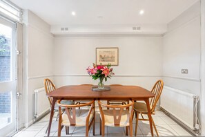 A dining room with a wooden table, six chairs, a vase of flowers, a framed picture on the wall, and a window with a view of greenery. - Large Chelsea Home With Private Garden (London)