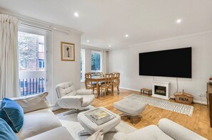 A living room with a sofa, ottoman, TV, fireplace, dining table, chairs, and a view through the window. - Chelsea Townhouse Garden Roof Terrace Pet Friendly (London)