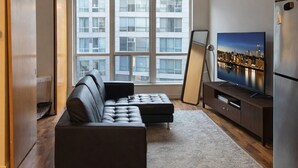 A modern living room with a sectional sofa, a flat-screen TV, a large window, and a cityscape view. - Modern Loft 1BD/1BA in King West (Toronto)