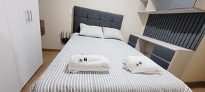 Een opgemaakt bed met streepjesbeddengoed en kussens, een nachtkastje met een lamp en een raam met jaloezieën.