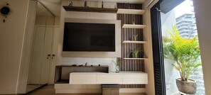 Een moderne woonkamer met een flatscreen televisie aan de wand, een inbouwkast en een grote potplant bij het raam.