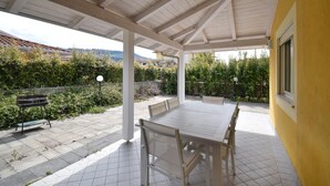 Un patio al aire libre con una mesa y sillas, rodeado de un jardín y una pared amarilla.